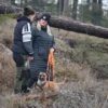 Två personer samtalar i skogsterräng med hund i sele och långlina vid sidan.