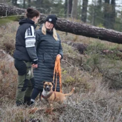 Två personer samtalar i skogsterräng med hund i sele och långlina vid sidan.